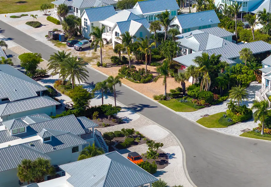Aerial view of a peaceful coastal neighborhood with blue-roofed homes, palm trees, landscaped yards, curving streets, and parked cars.