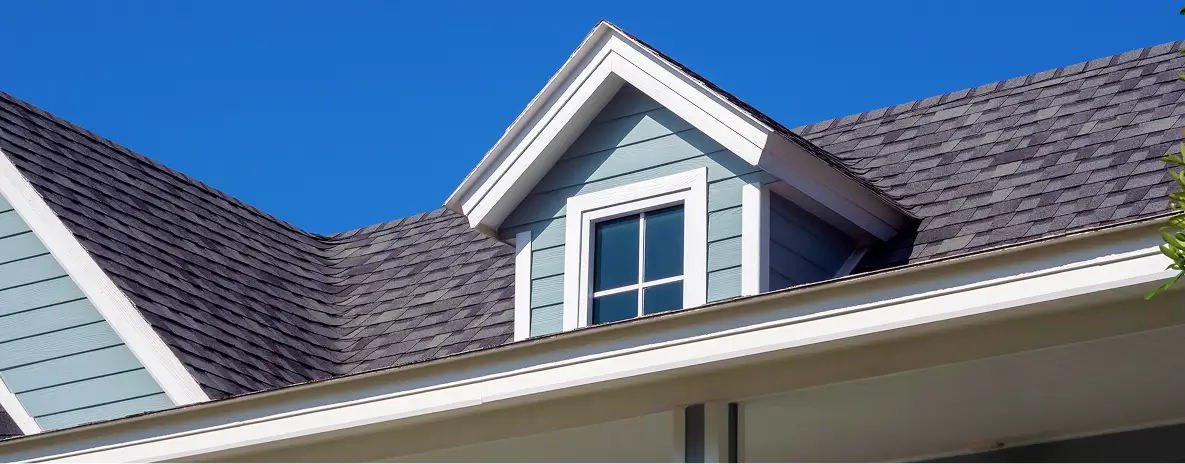 Close-up of a charming gray house roof and dormer window with white trim under a vivid blue sky, creating a bright, welcoming mood.