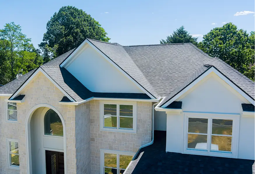 Sunlit stone-and-white suburban house with gray shingle roofs, arched entry, large windows, leafy trees, and a clear blue sky.