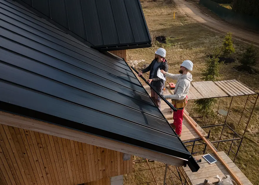 Two workers in white hard hats install dark metal roofing on a wooden building, balancing on scaffolding in a sunny rural setting.