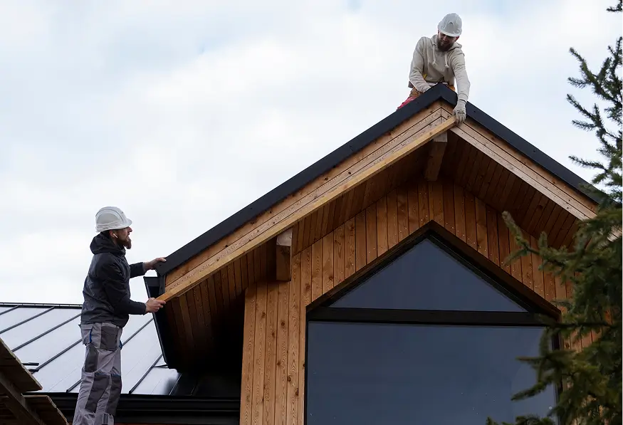 Two roofers in safety gear install dark trim on a wooden house roof, one perched at the peak and one working below under a cloudy sky.