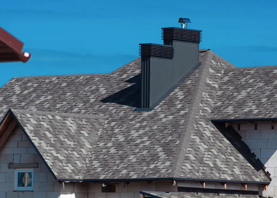 Close-up of a weathered shingle roof with two tall metal chimneys against a clear blue sky, conveying rustic architectural detail.