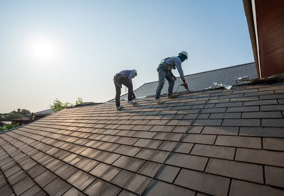 Two roofers in safety gear work on a slate-tiled rooftop under bright sun, conveying focused teamwork and professional home maintenance.